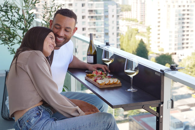 A couple sitting close together and smiling while enjoying snacks and wine on the Zenzo Balcony Bar. The bar is set up on a glass balcony overlooking a city skyline with a bottle of wine, glasses, and a wooden snack platter on the bar.