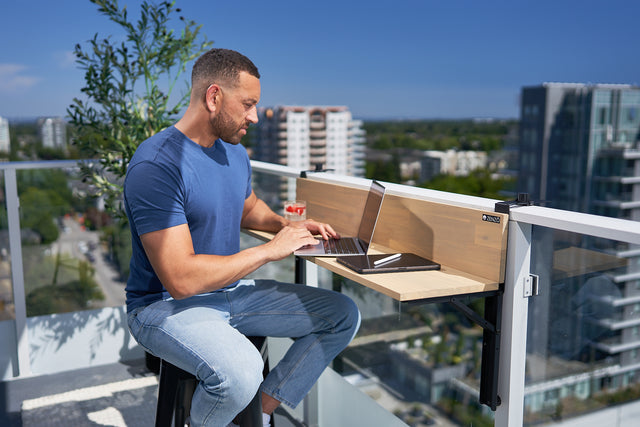 A man using a laptop on the Zenzo Balcony Bar, set up on a glass balcony with a cityscape view. The bar holds a glass of juice, notebook, and pen, showing its use as a functional workspace in an outdoor setting.