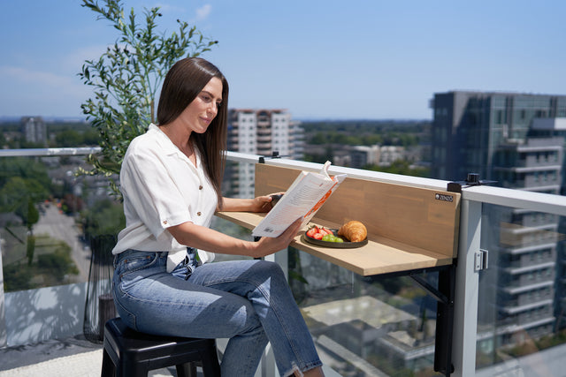 A woman reading a book while seated at a Zenzo balcony table, with a plate of fruit and a croissant, overlooking an urban skyline.