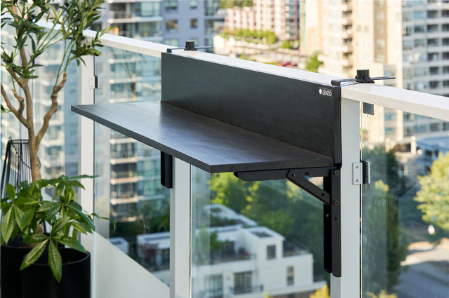 Zenzo Balcony Bar in black wood finish installed on a glass balcony railing. The bar is empty, highlighting its modern design with a city view and potted greenery in the background.