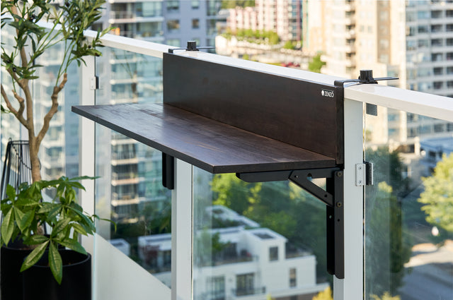 Zenzo Balcony Bar in a dark oak wood finish installed on a glass balcony railing. The bar is empty, displaying its elegant design with a view of city buildings and potted plants in the background.