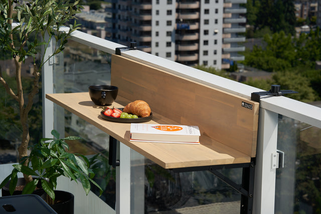 Zenzo Balcony Bar in birch finish set up on a glass balcony railing. The bar holds a black coffee mug, a plate with fruit and a croissant, and an open book, creating a relaxing spot with a view of the city and surrounding greenery.