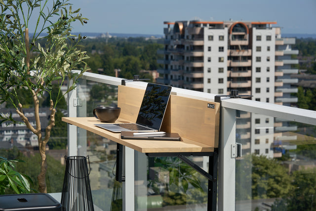 Zenzo Balcony Bar in birch finish set up as a workspace on a glass balcony railing. The bar holds a laptop, a black bowl, a notebook, and a pen, with a city skyline and greenery in the background.