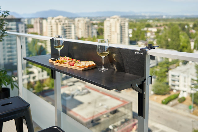 Zenzo Balcony Bar set up on a glass balcony, featuring two glasses of white wine and a wooden platter with snacks. The bar overlooks a scenic cityscape with buildings and greenery in the background.